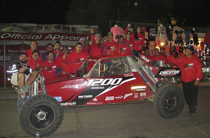 Race team celebrating beside their buggy at the 2016 Baja 1000 finish line at night
