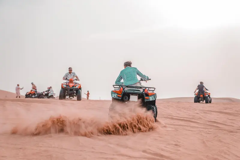 Riders on ATVs kicking up sand on a dune in bright sunlight