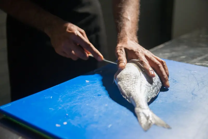 Cook cleaning a whole fresh fish with a knife on a blue cutting board