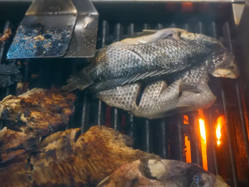 Whole fresh fish cooking on a charcoal grill next to a metal spatula