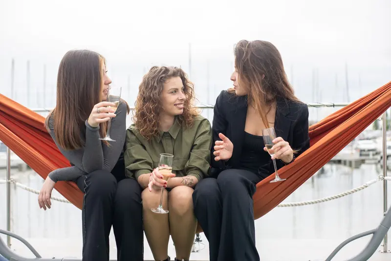 Three women sitting in a hammock with glasses of wine at a marina