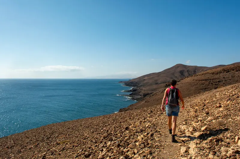 A hiker with a backpack walking a dry rocky trail above the blue sea on a clear day