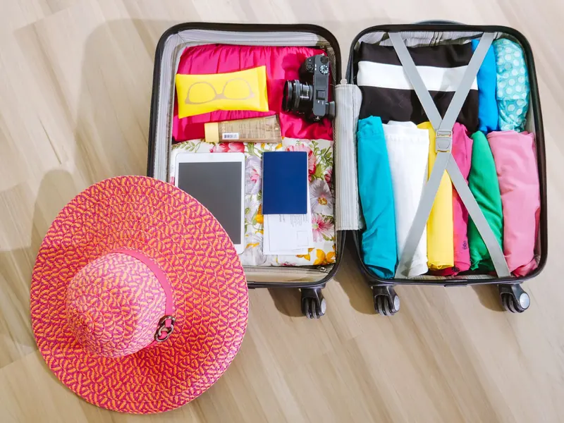 Maleta abierta con ropa de colores doblada, lentes, pasaporte y un sombrero rosa junto a ella