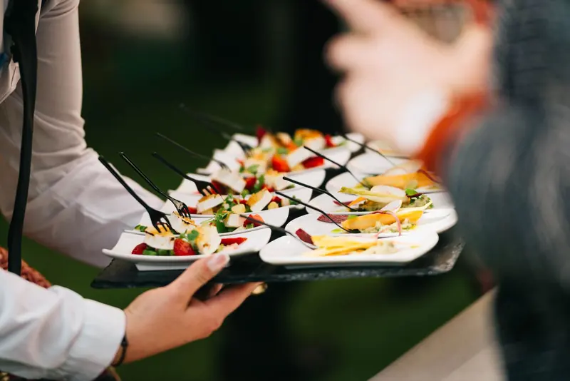 Server carrying a tray of small gourmet bites on white dishes at an evening event