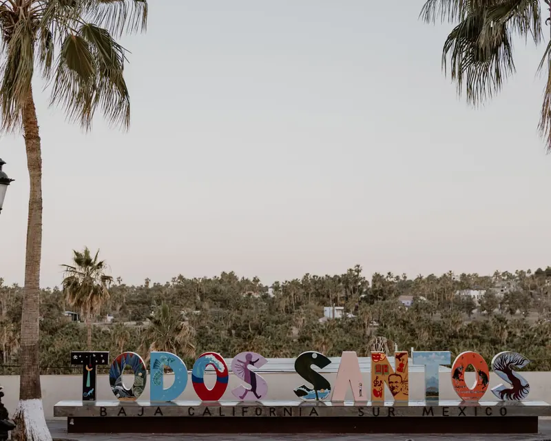 The colorful Todos Santos town sign with palm trees and the green valley behind it