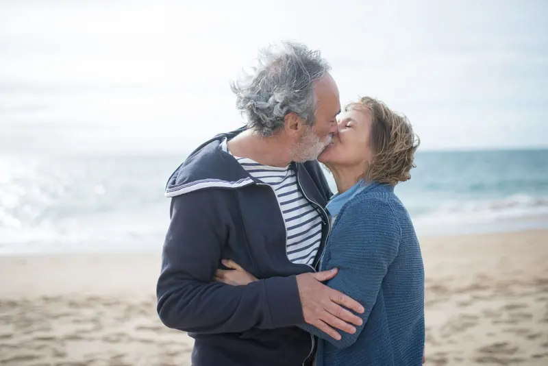 An older couple kissing on a beach with the sea behind them