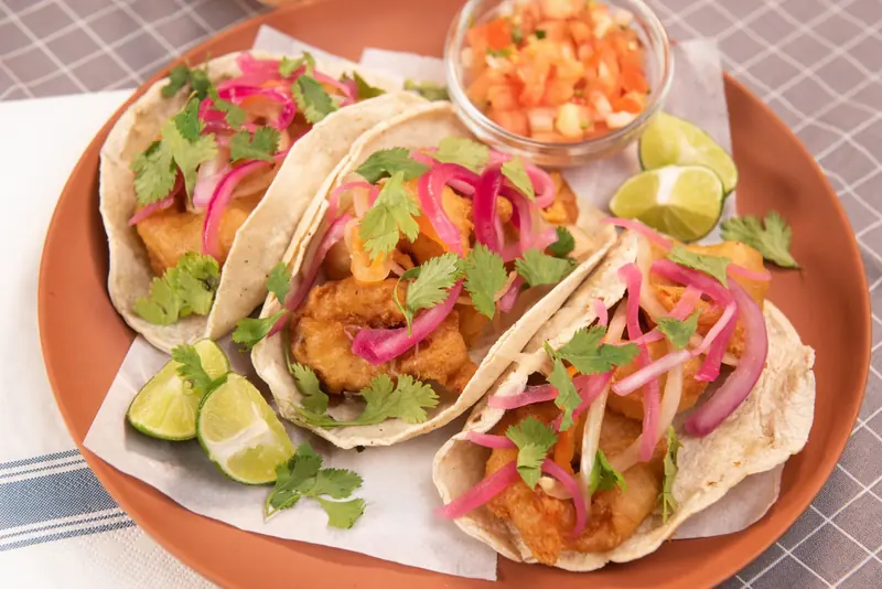Battered fish tacos topped with pickled red onion and cilantro, with lime wedges and salsa