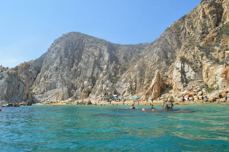 Agua turquesa y transparente al pie de los cerros de Finisterra, con pelícanos sobre una roca y gente en la arena